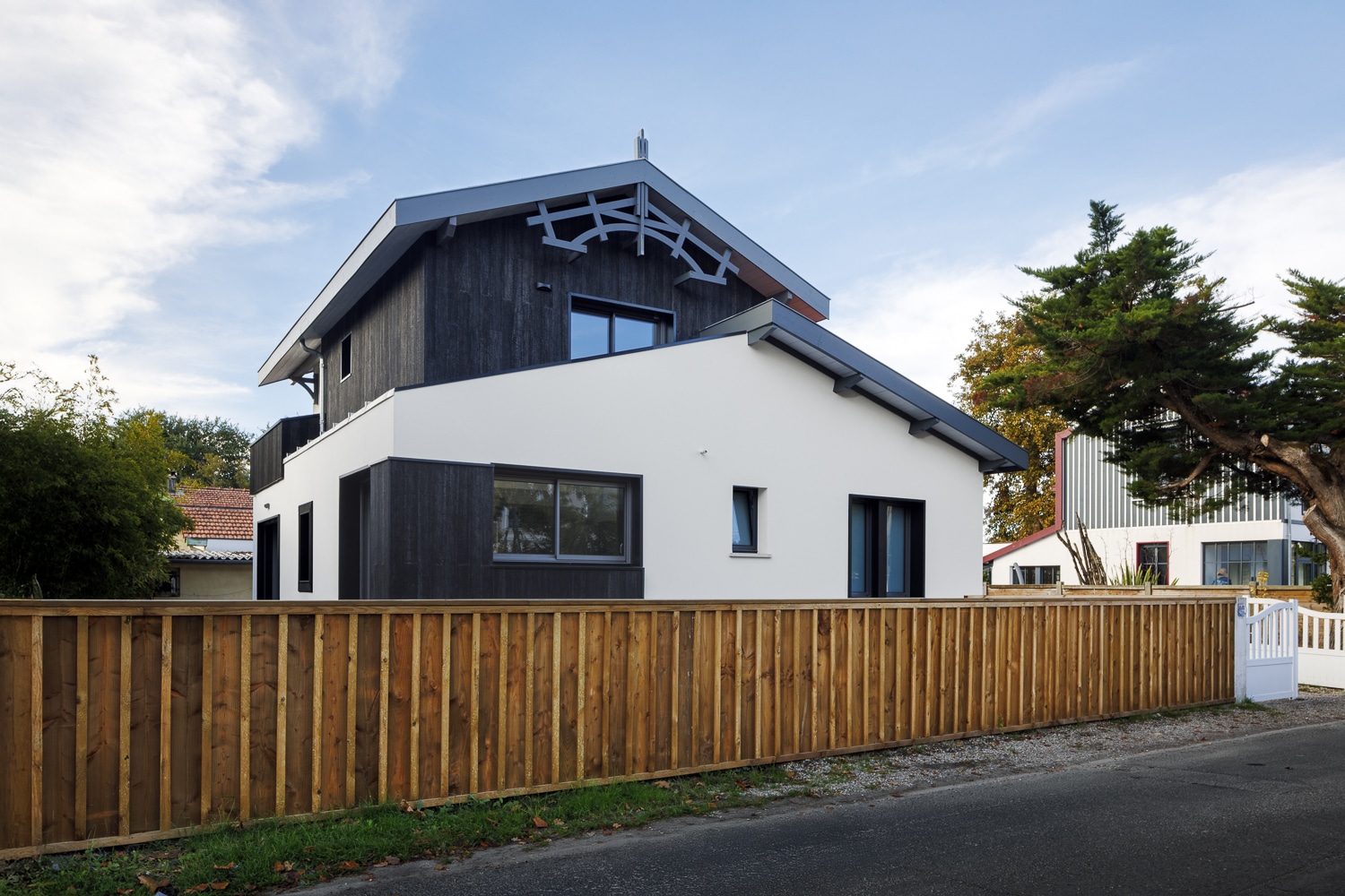 Maison moderne blanc et bois noir, clôture en bois, sous un ciel bleu parsemé de nuages.