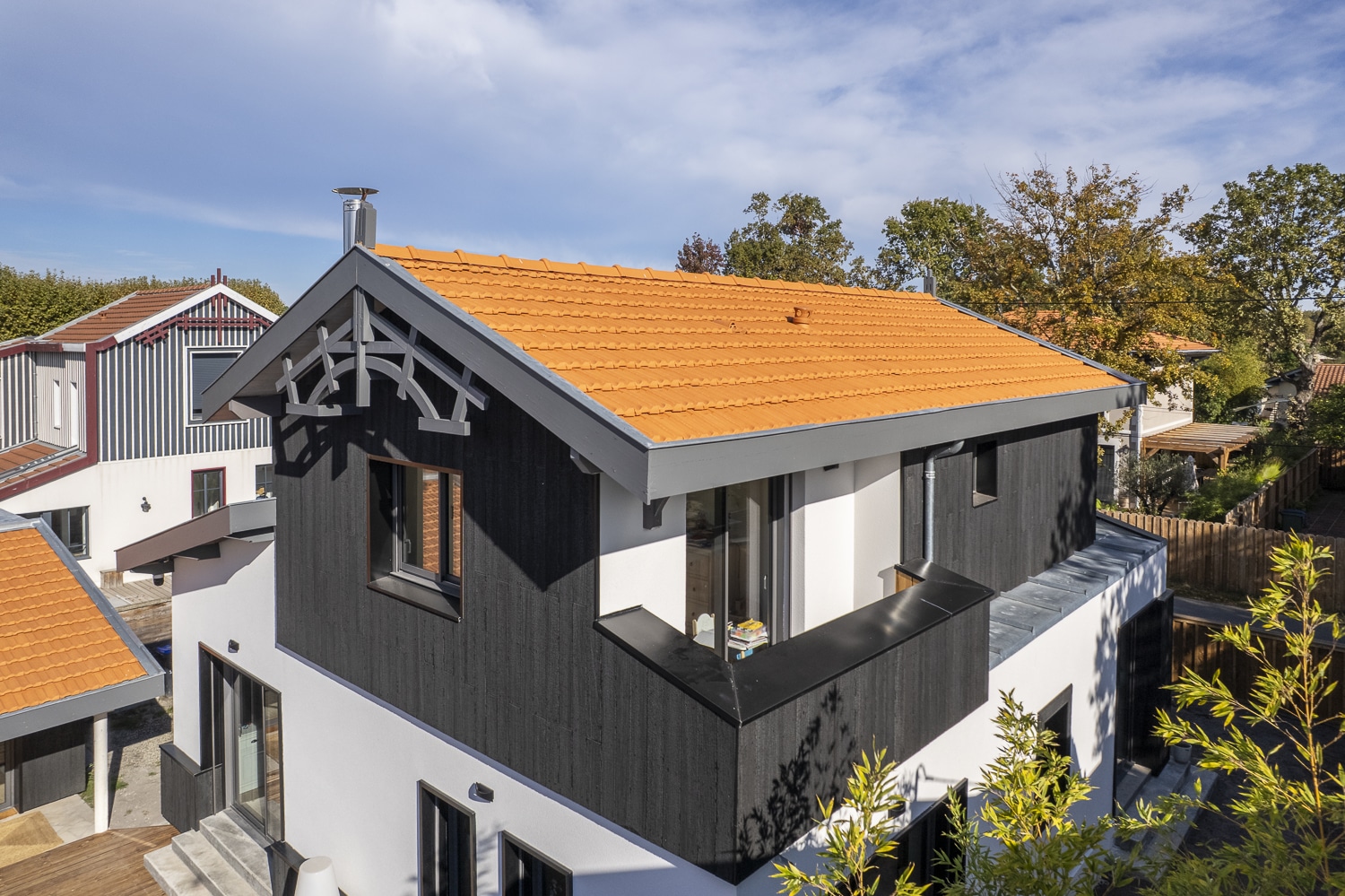 Maison moderne avec bardage bois noir, toiture tuiles terre cuite et colombages décoratifs, vue extérieure ensoleillée.