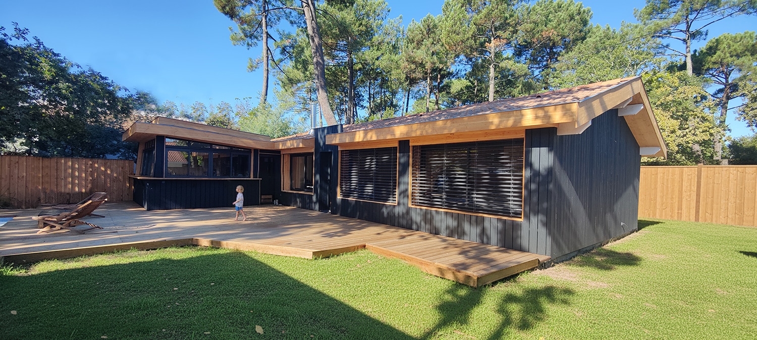Maison moderne noire et bois, terrasse en bois claire. Un enfant marche sur le deck, jardin verdoyant et pins sous un ciel bleu.