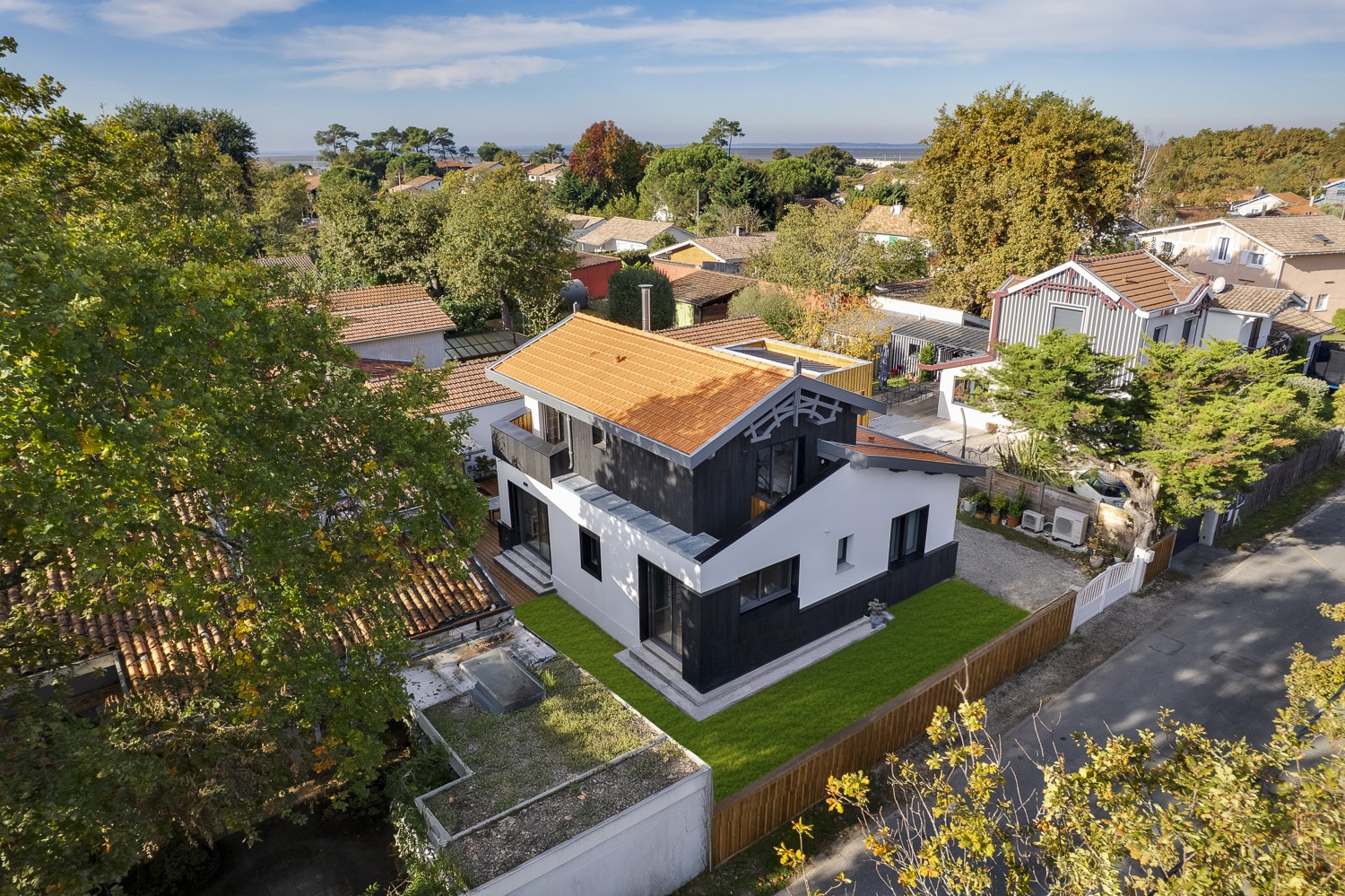 Vue aérienne maison moderne noir et blanc toit tuiles orange, entourée d'arbres et de toits traditionnels.