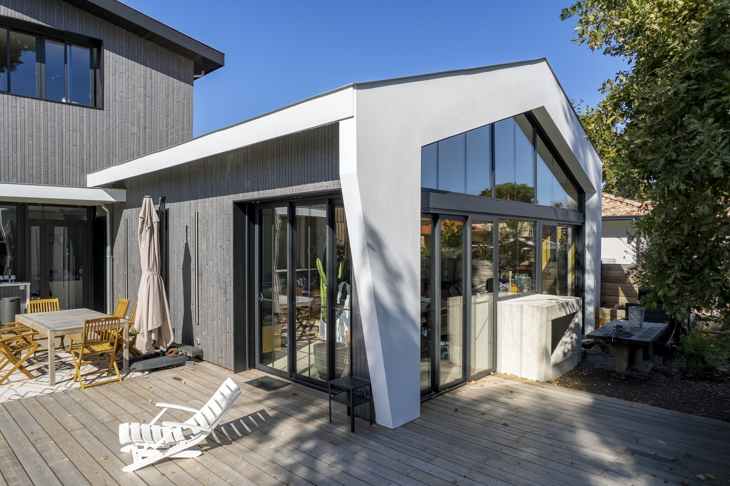 Terrasse en bois d'une maison moderne avec bardage gris, grande baie vitrée et structure blanche inclinée.