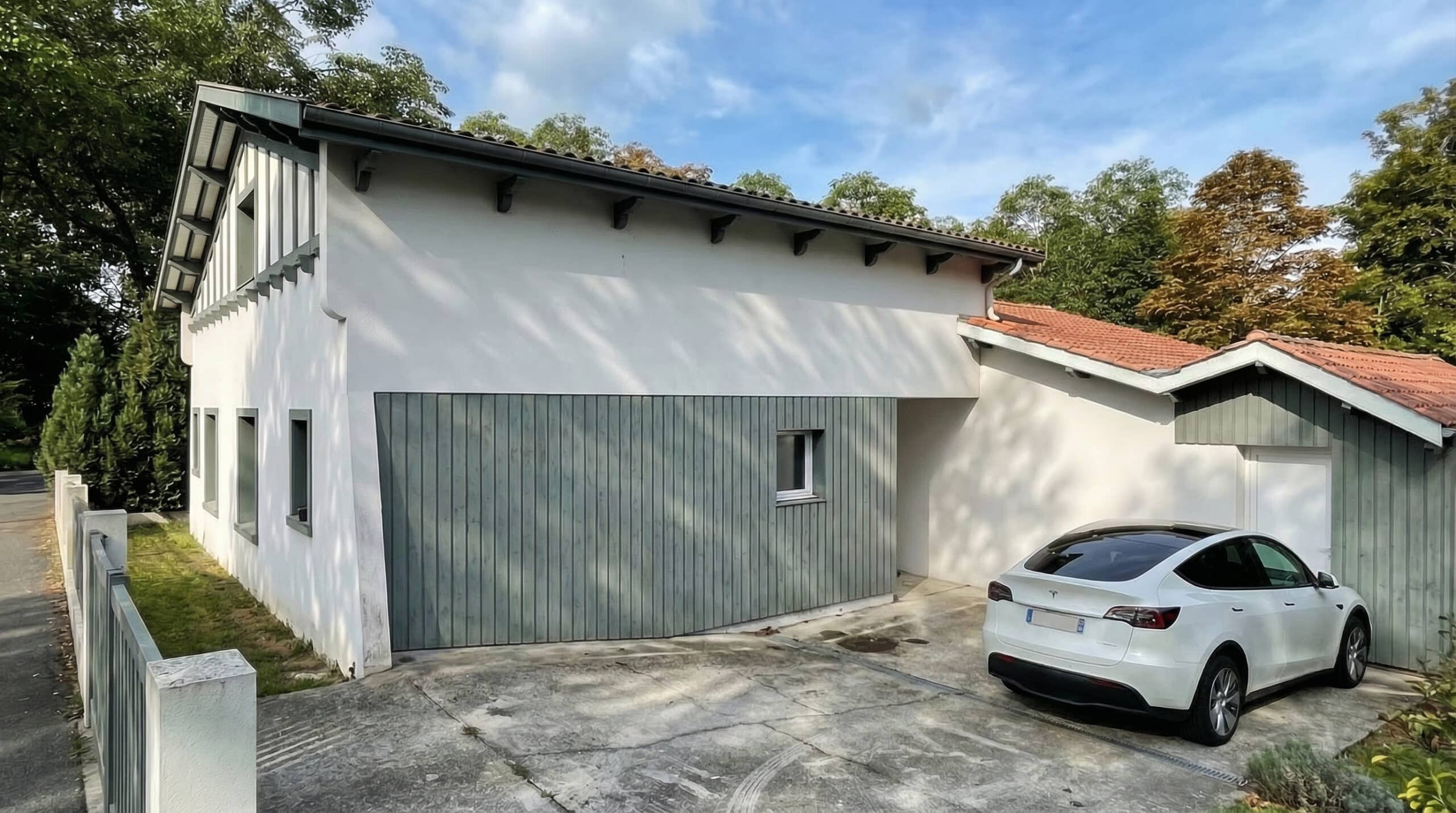 Maison contemporaine blanche avec bardage bois gris et une Tesla blanche garée dans l'allée ensoleillée.