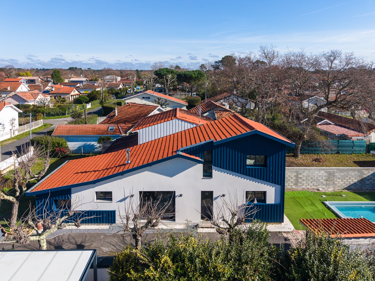 Maison moderne bleue et blanche au toit orange avec piscine, vue aérienne dans un quartier résidentiel ensoleillé.