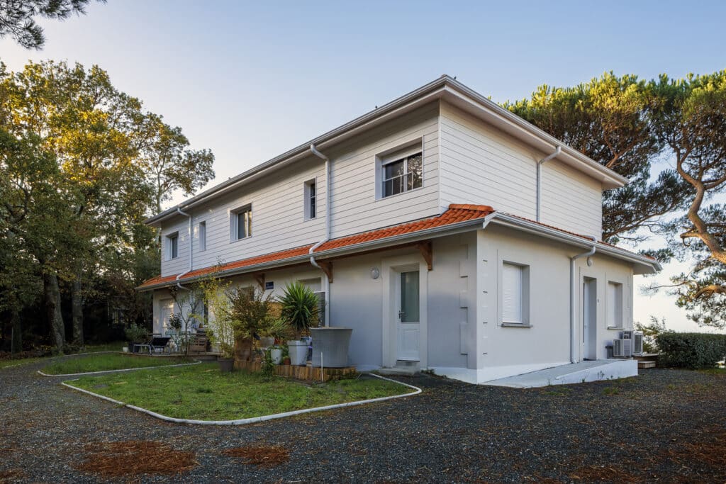 Maison jumelée moderne à deux étages, revêtement blanc et toit en tuiles rouges, entourée d'arbres, avec allée en gravier.