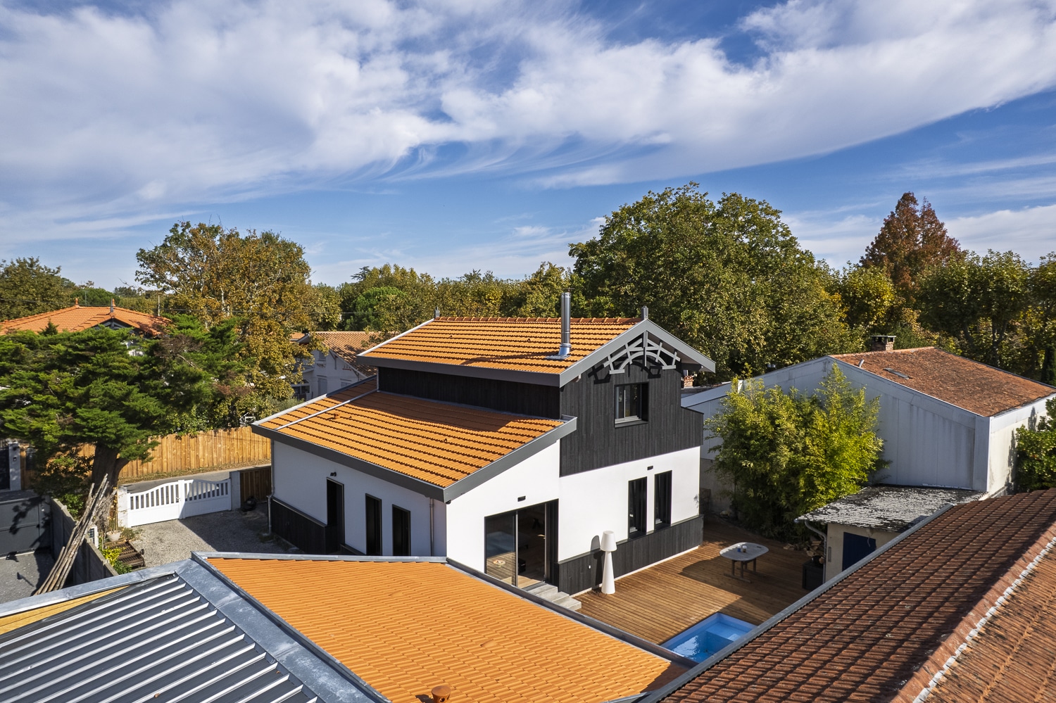 Maison moderne blanc et noir avec toit en tuiles orange vif, terrasse en bois et petite piscine, sous un ciel bleu parsemé de nuages.