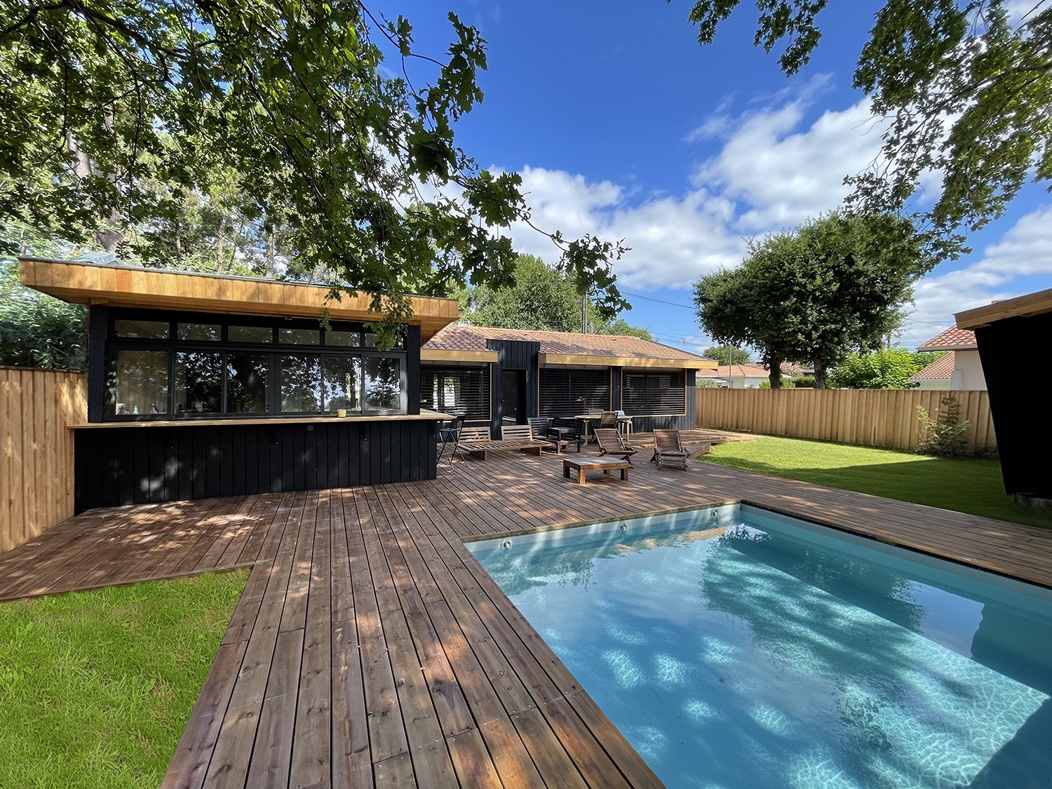 Luxueux espace extérieur avec piscine, vaste terrasse en bois et bâtiment moderne noir sous un ciel bleu. Pelouse verte et arbres ombrageux.
