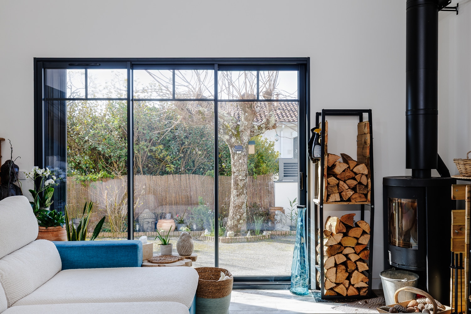 Salon avec baie vitrée noire sur jardin, poêle à bois moderne et étagère de bûches. Ambiance chaleureuse et lumineuse.