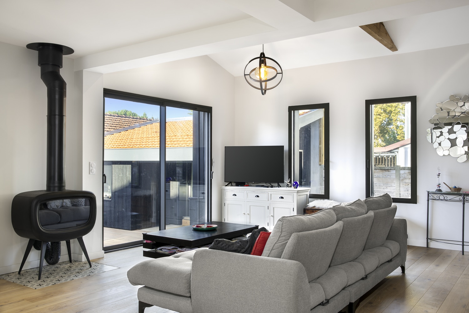 Salon moderne clair avec poêle à bois noir design, canapé gris, meuble TV blanc et grandes baies vitrées donnant sur un extérieur ensoleillé.