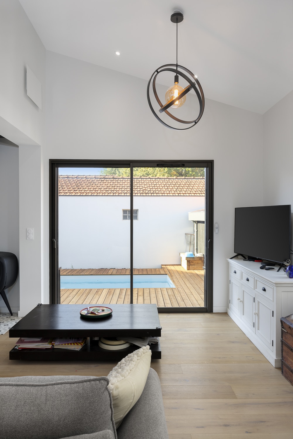 Salon moderne avec plafond cathédrale, suspension design noire, plancher bois clair et baie vitrée donnant sur une terrasse bois et piscine.