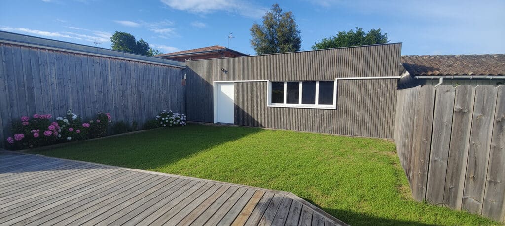 Cour extérieure moderne avec terrasse en bois gris, pelouse verte et bâtiment avec bardage bois foncé, porte blanche et fenêtre.