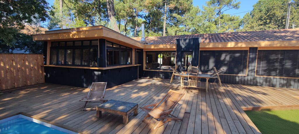 Architecture moderne : maison noire et bois avec grande terrasse en bois, piscine et mobilier extérieur. Entourée d'une pinède luxuriante.