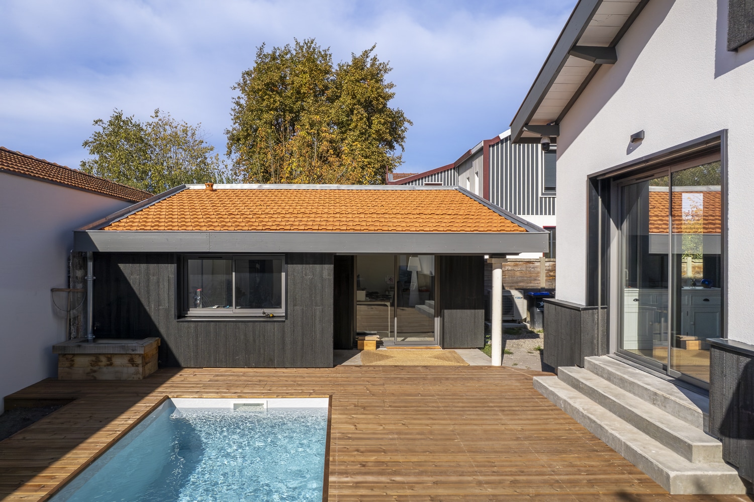 Terrasse en bois moderne avec piscine, pavillon noir à toit tuile orange, et façade blanche sous ciel bleu.