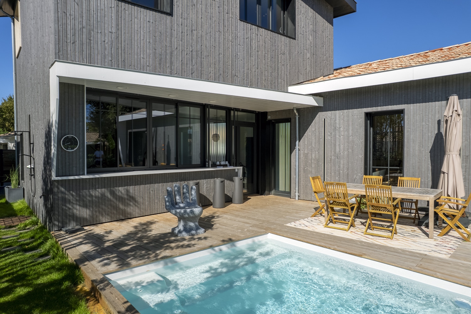 Terrasse en bois moderne avec petite piscine, salon de jardin bois et sculpture main, bardage gris.