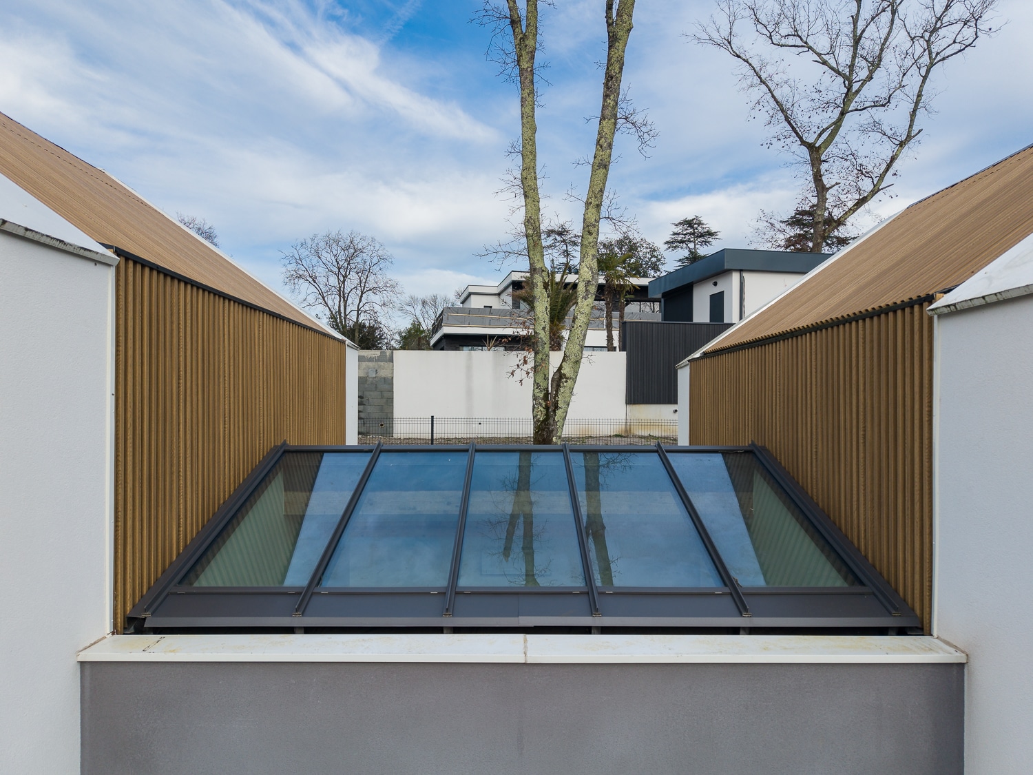 Verrière moderne entre deux pans de murs en bardage métallique marron, avec arbres et ciel bleu en arrière-plan.