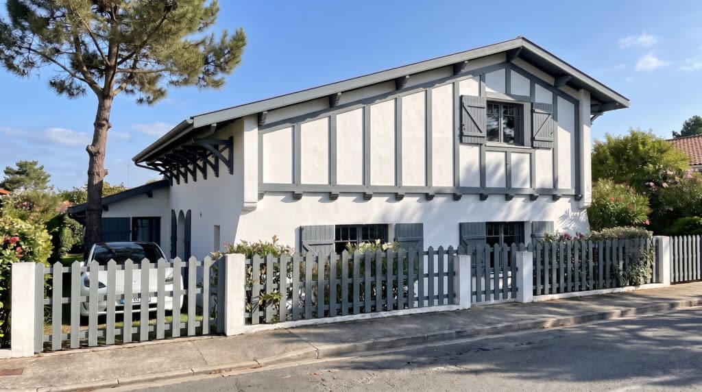 Villa néo-basque blanche avec colombages gris, clôture assortie et voiture électrique devant.