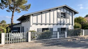 Villa néo-basque blanche avec colombages gris, clôture assortie et voiture électrique devant.