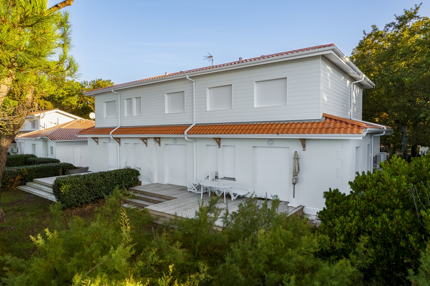 Villas jumelées blanches avec toits en tuiles orange et volets roulants fermés, terrasse en bois et verdure luxuriante.