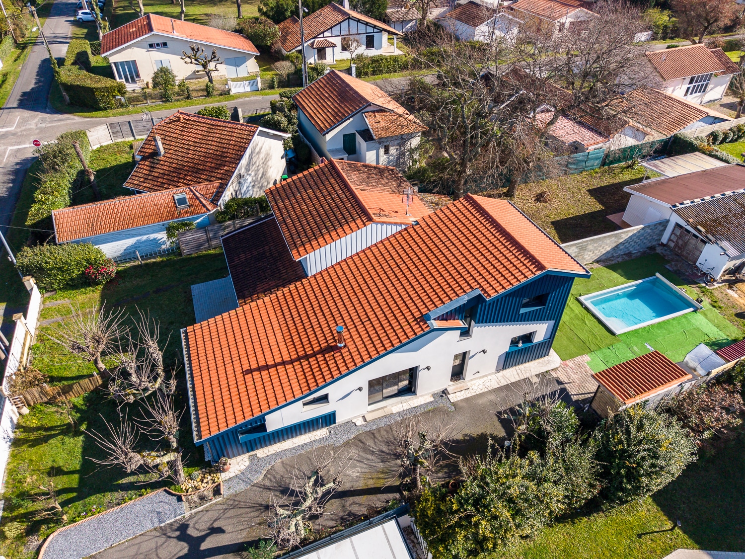 Vue aérienne d'une maison moderne à toit de tuiles orange et piscine dans un quartier résidentiel arboré.