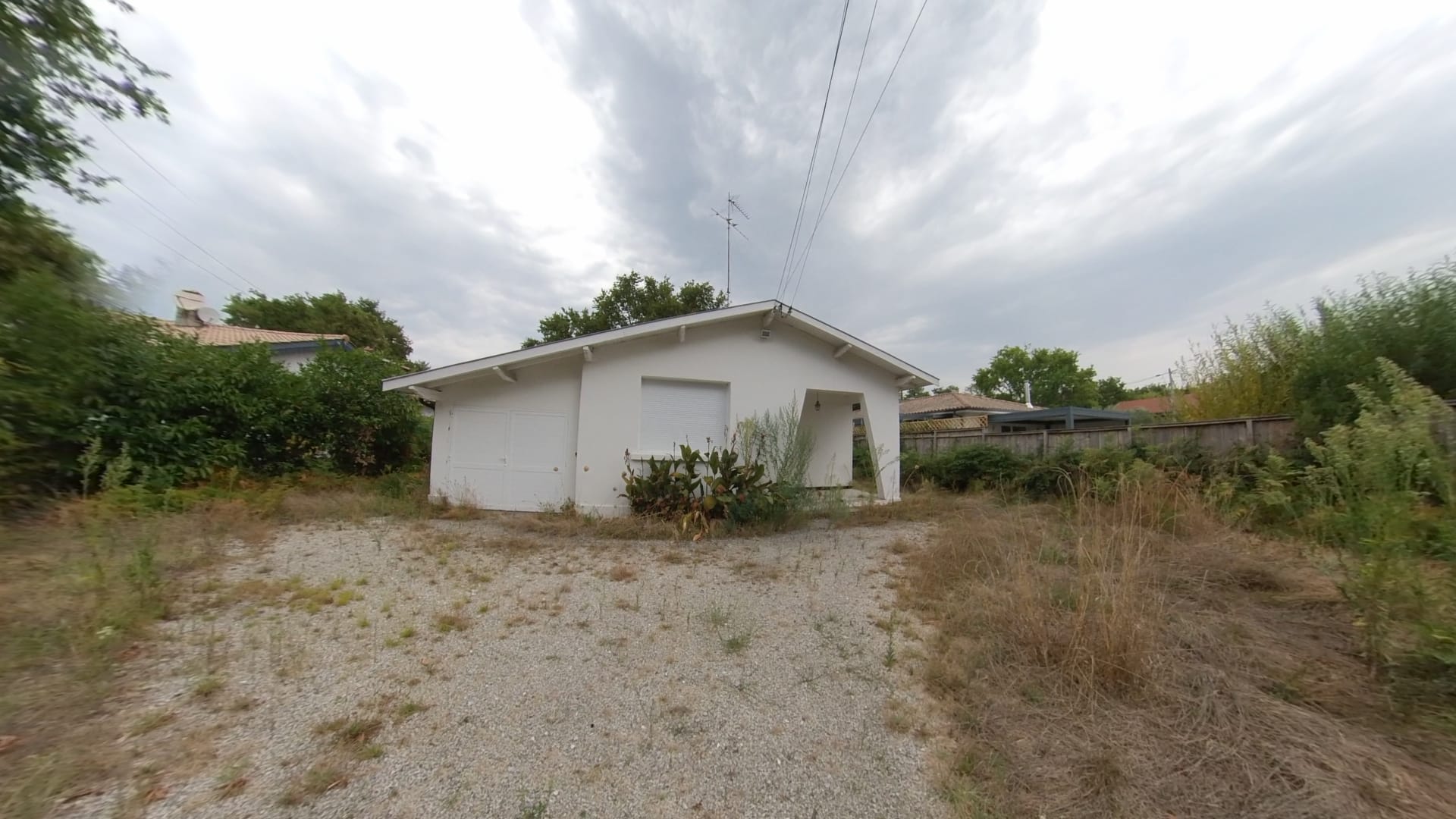 Maison blanche de plain-pied avec jardin en friche, allée en gravier et ciel nuageux menaçant.