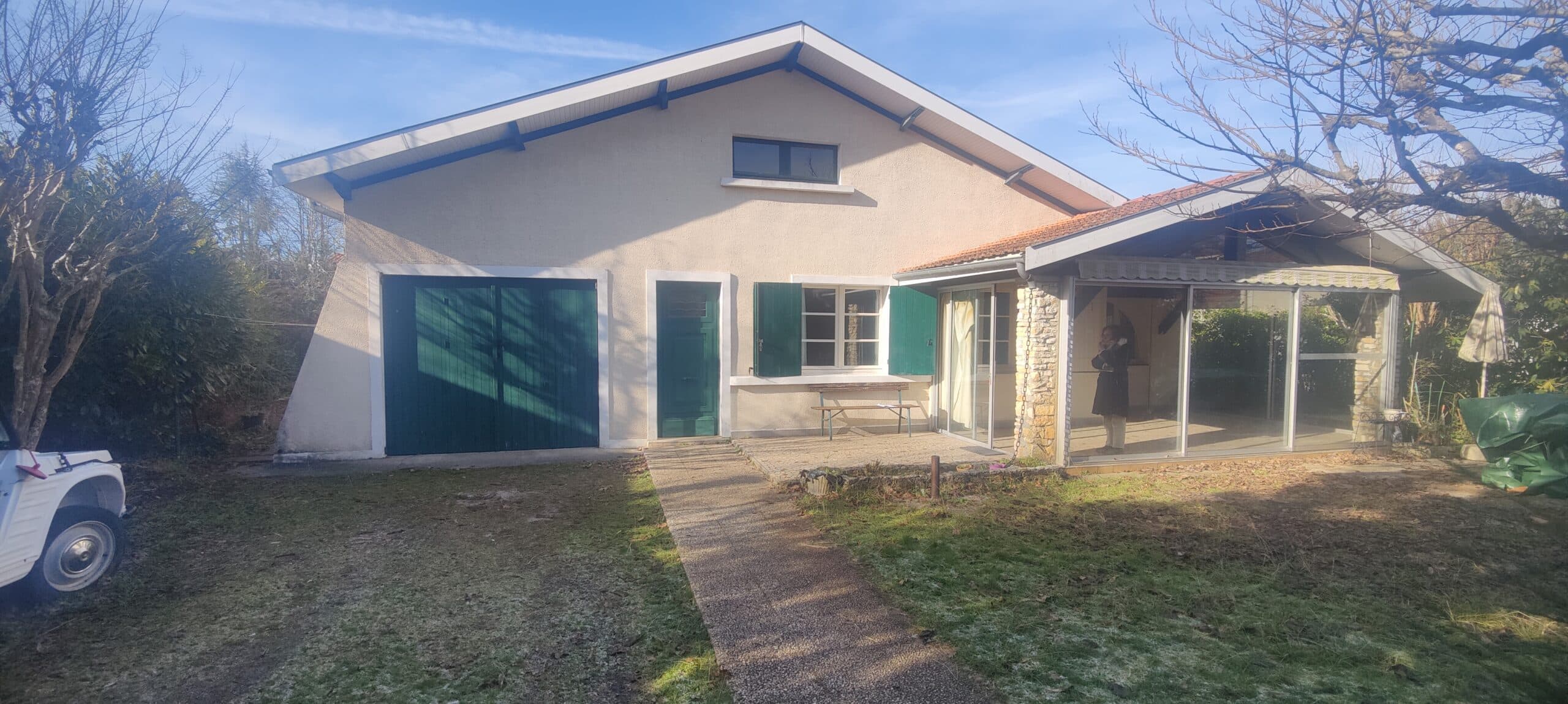 Maison à façade beige avec garage et volets verts, véranda vitrée latérale et voiture vintage sur un terrain herbeux.