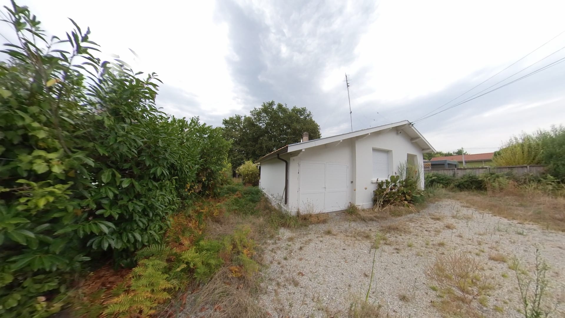 Petite maison blanche avec garage dans un jardin sauvage sous un ciel gris et nuageux.